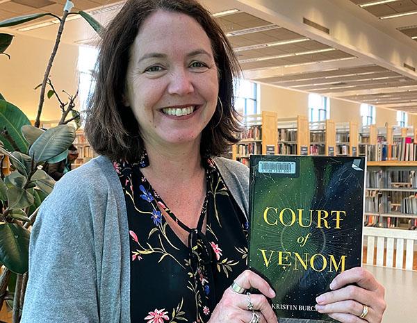Author looking at the camera holding up a library copy of their book, Court of Venom.