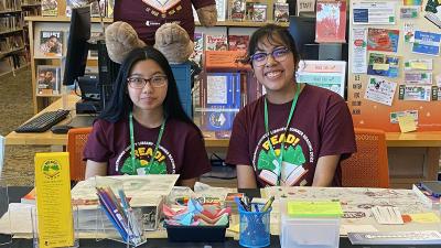 Two teen volunteers seated at the Summer Reading table inside the library