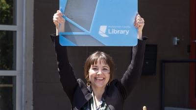 Annie Lewis, new interim director standing outside with her arms up holding a giant library card above her head,