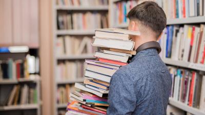 Person inside a library, holding a large stack of books with their back turned.