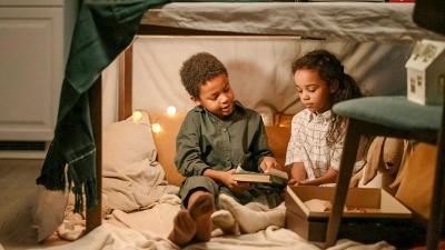 Two children read a book in a blanket fort with pillows and lights set up underneath a table.