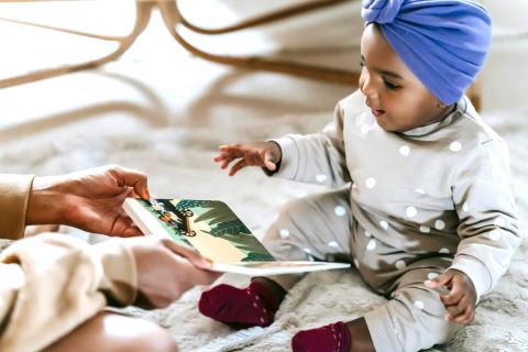 a brown skinned baby sitting on the floor wearing a bonnet looking at a board book