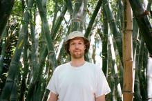 Photograph of a man wearing a white t-shirt and bucket hat in front of a bamboo grove