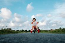 Photo of a toddler riding a red tricycle with a clouds and blue sky background.
