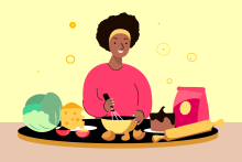A brown skinned woman in a pink shirt smiling and preparing food surrounded by ingredients