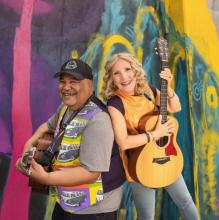 photograph of two musicians holding instruments, smiling back to back in front of a colorful background