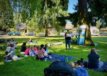 Families sit on blankets in the park as a librarian reads a book aloud.