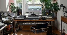 Photo of a setup for creating music at a wooden desk, including a piano and monitor with music editing software.