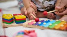 The hands of a baby and an adult play with brightly colored toys including a xylophone and building bricks.