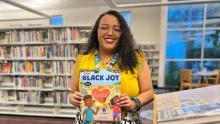Photo of Isabel Villareal Stewart smiling and holding a Black Family Storytime book titled Black Joy.