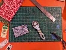 A green cutting mat is shown with several tools used for sewing including a rotary cutter, fabric scissors, a pack of needles, a ruler and fabrics.