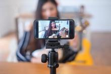 A photograph of an Asian woman using a smartphone to film herself play guitar