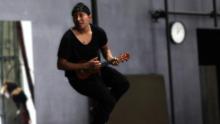 Photo of Fyre Daway, aka Gigs, sitting on a stool while playing an instrument.