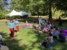 Two women in blue and red read stories to a group of adults and young children in a park.
