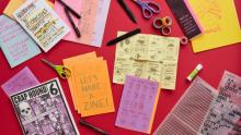 Photo of several different papers with different words and designs laid out on a red surface with glue sticks, scissors, and pens around them.