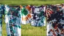 Photo of several tie dye shirts in many colors drying on a clothesline outside.