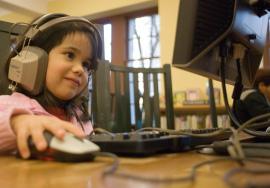 Niña sonriente con audífonos usando un ratón para controlar una computadora personal en una biblioteca.