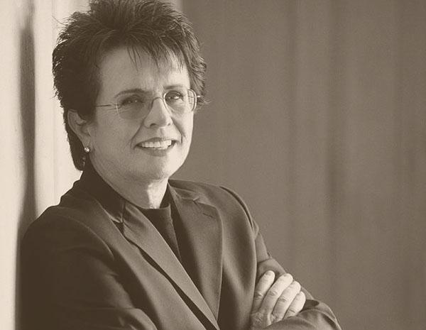 Billie Jean King leaning against wall with arms crossed and smiling at camera.