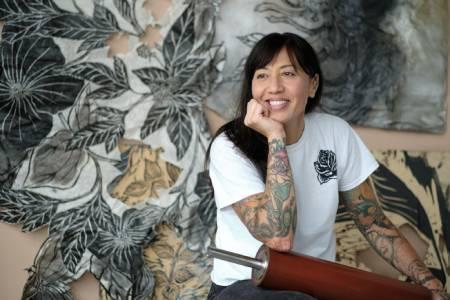 Artist Kanani Miyamoto sits in front of a wall print featuring dark leaves.