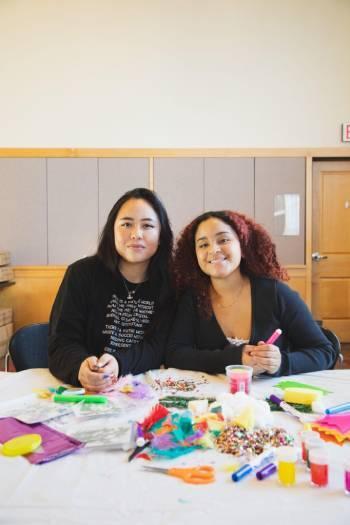 Artists Lillyanne Pham and Paola De La Cruz are seated, smiling at the camera, with art supplies on the table in front of them.