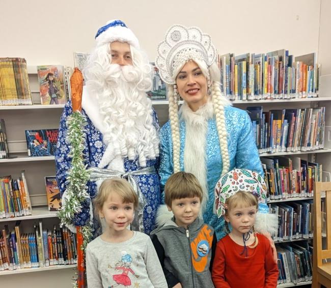 Дед Мороз и Снегурочка стоят за спинами троих детей, отмечающих Новый год в библиотеке