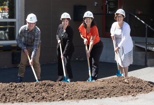 Four people standing behind a pile of dirt, wearing hard hats, standing with shovels and smiling.
