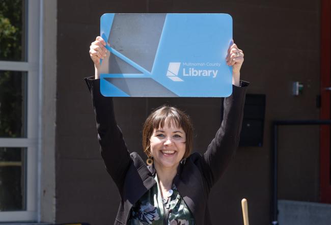 Annie Lewis standing outside holding a giant library card above her head smiling at the camera