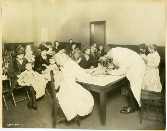 An old photograph of a baby clinic inside a library, people sitting in chairs, a woman with her baby at the front of the room, the doctor is inspecting the baby on a table.