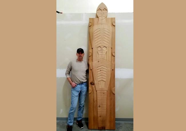 Artist standing against a wall with a large wooden sculpture next to him