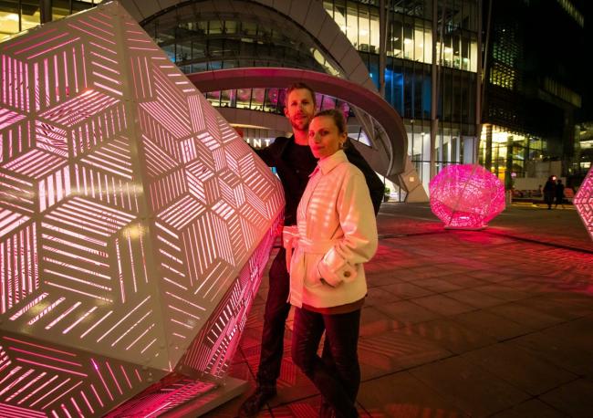 Two people, a man and a woman standing next to an octagon type large sculpture at night time