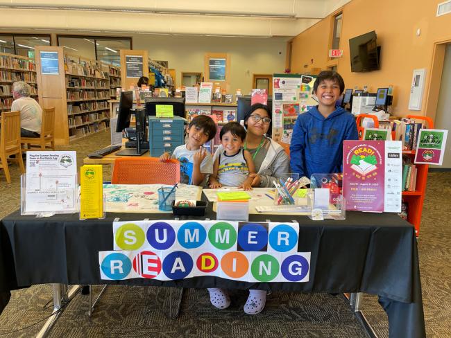 Summer Reading volunteer with three kids sitting behind a table with a banner that says Summer Reading