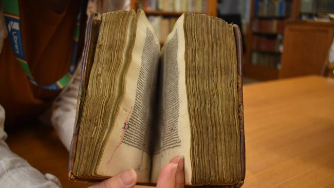 hands hold open a medieval book with miniscule text in Latin and a worn leather binding